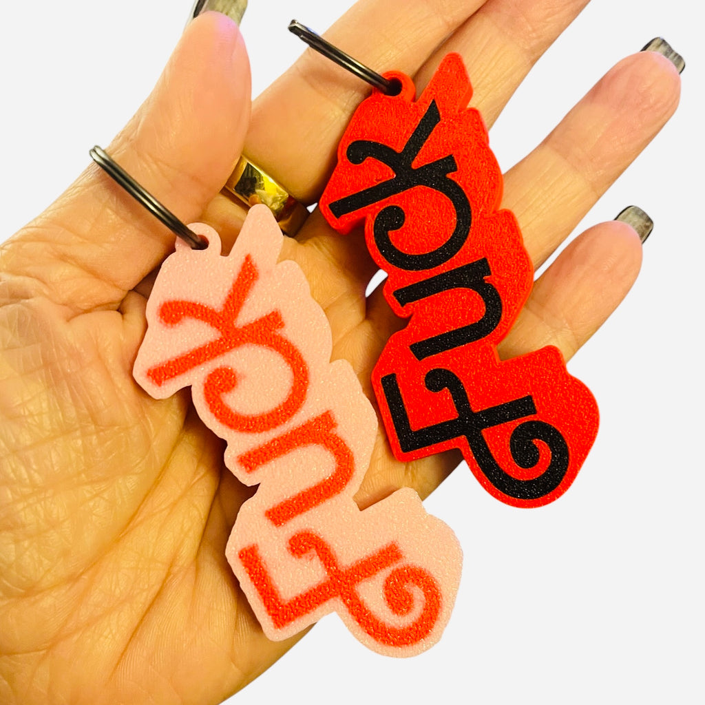 Two novelty keychains spelling 'FUCK' in stylized Barbie script, held in a hand. One keychain is light pink with red lettering, and the other is bright red with black lettering.
