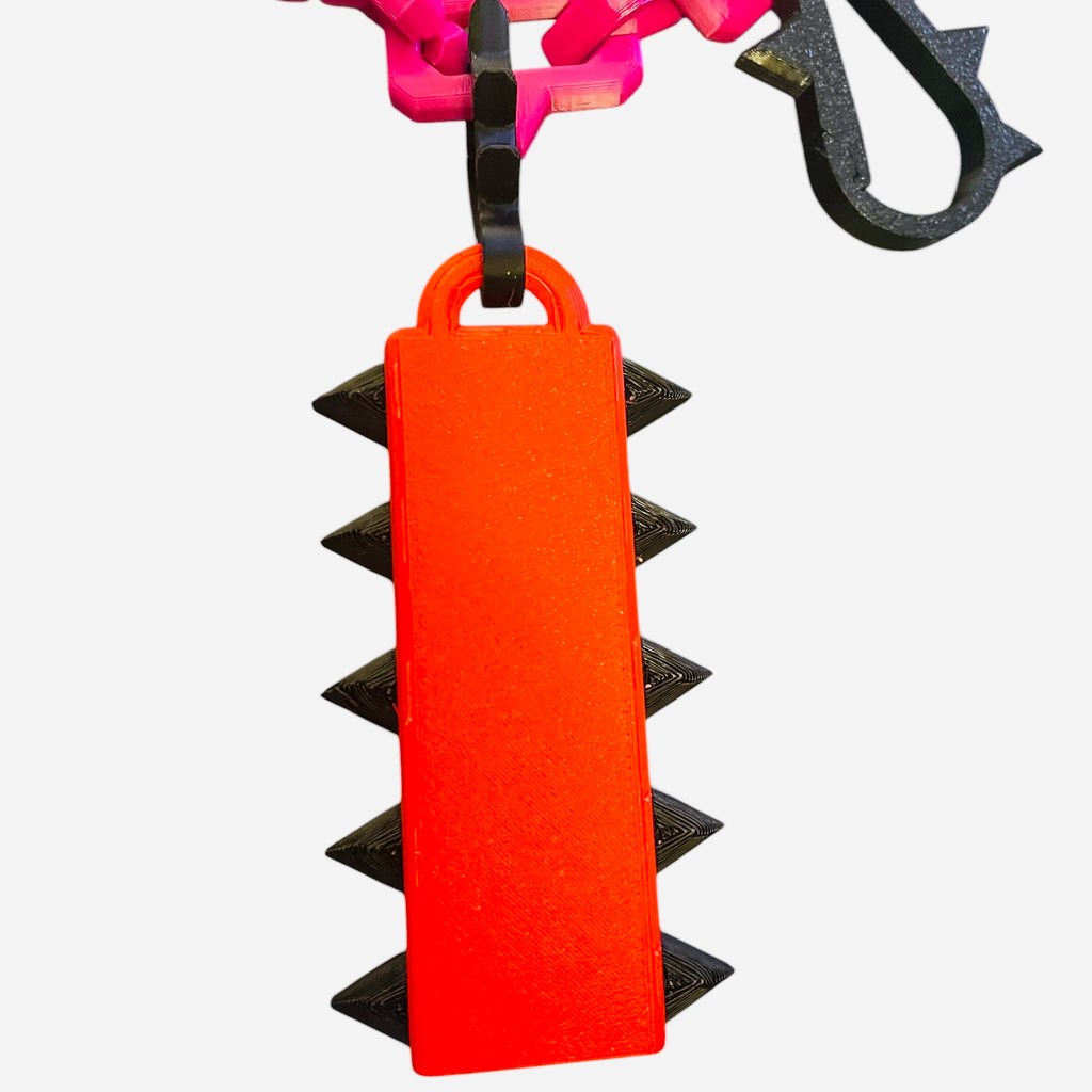 A bright red rectangular keychain or bag charm with black spikes along the sides, attached to a black ring and pink chain, shown against a plain white background.