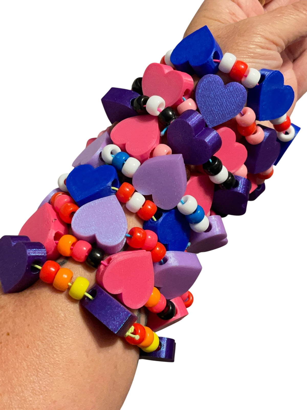A close-up of a person's wrist wearing a stack of colorful heart-shaped 3d printed beaded bracelets. The bracelets feature heart charms in pink, purple, and blue, alongside multicolored beads in red, orange, yellow, white, and black.