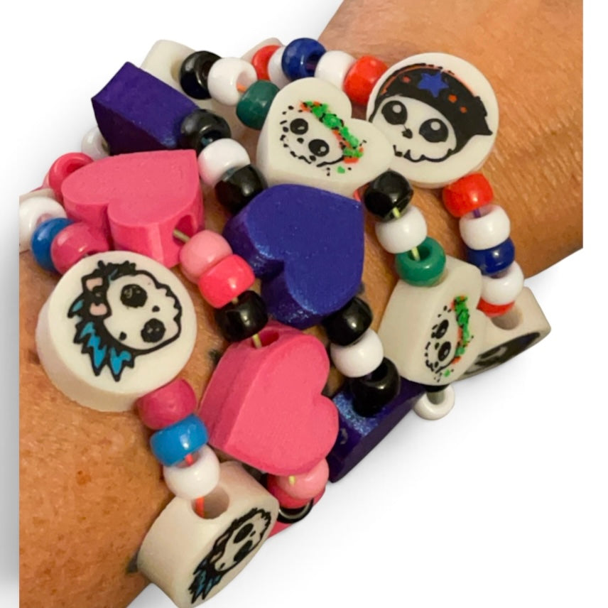A close-up of a person's wrist wearing a stack of colorful 3d printed beaded bracelets with heart, and round charms featuring skull designs.