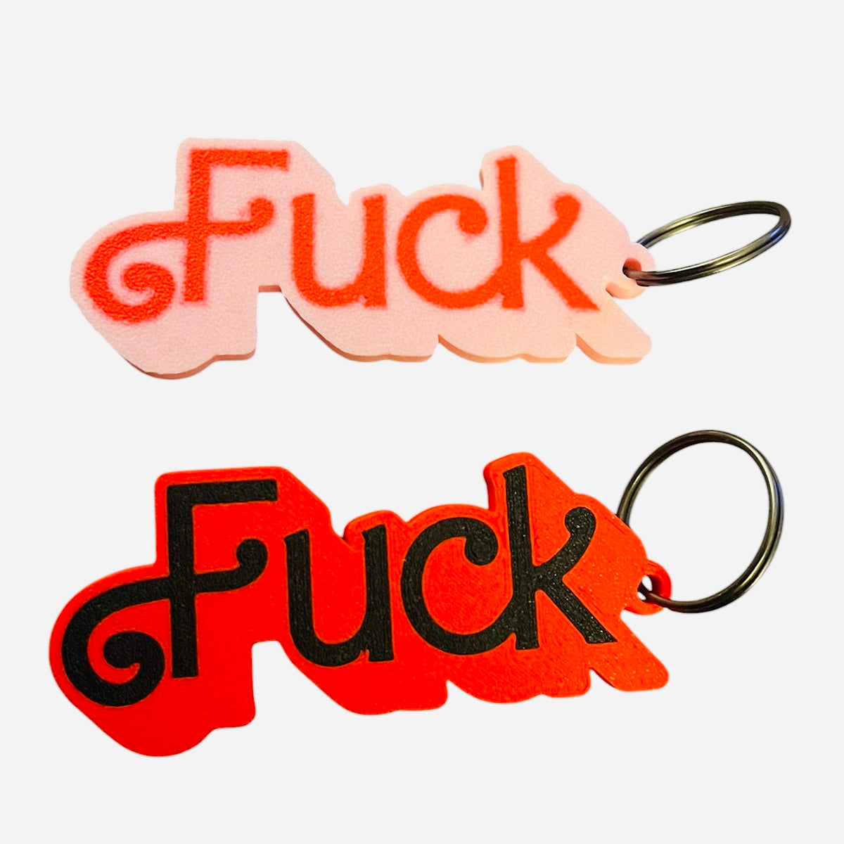 Two novelty keychains spelling 'Fuck' in stylized cursive Barbie lettering, one in light pink with red text and one in red with black text, both attached to black metal key rings on a white background.