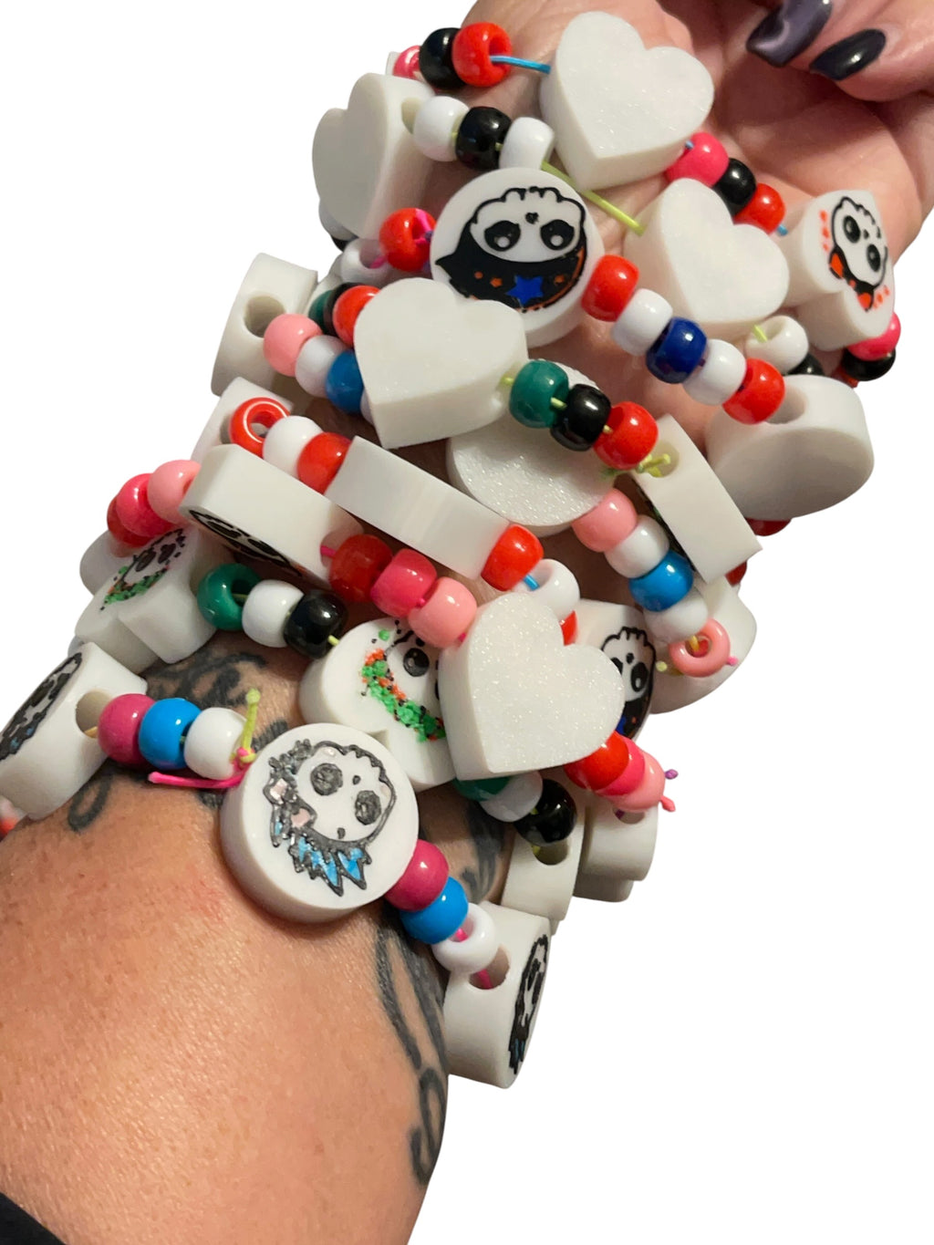 A close-up of a person's wrist wearing a stack of 3d printed beaded bracelets. The bracelets feature white heart-shaped charms, round white charms with black and colorful skull illustrations, and colorful pony beads in red, pink, blue, green, black, and white.