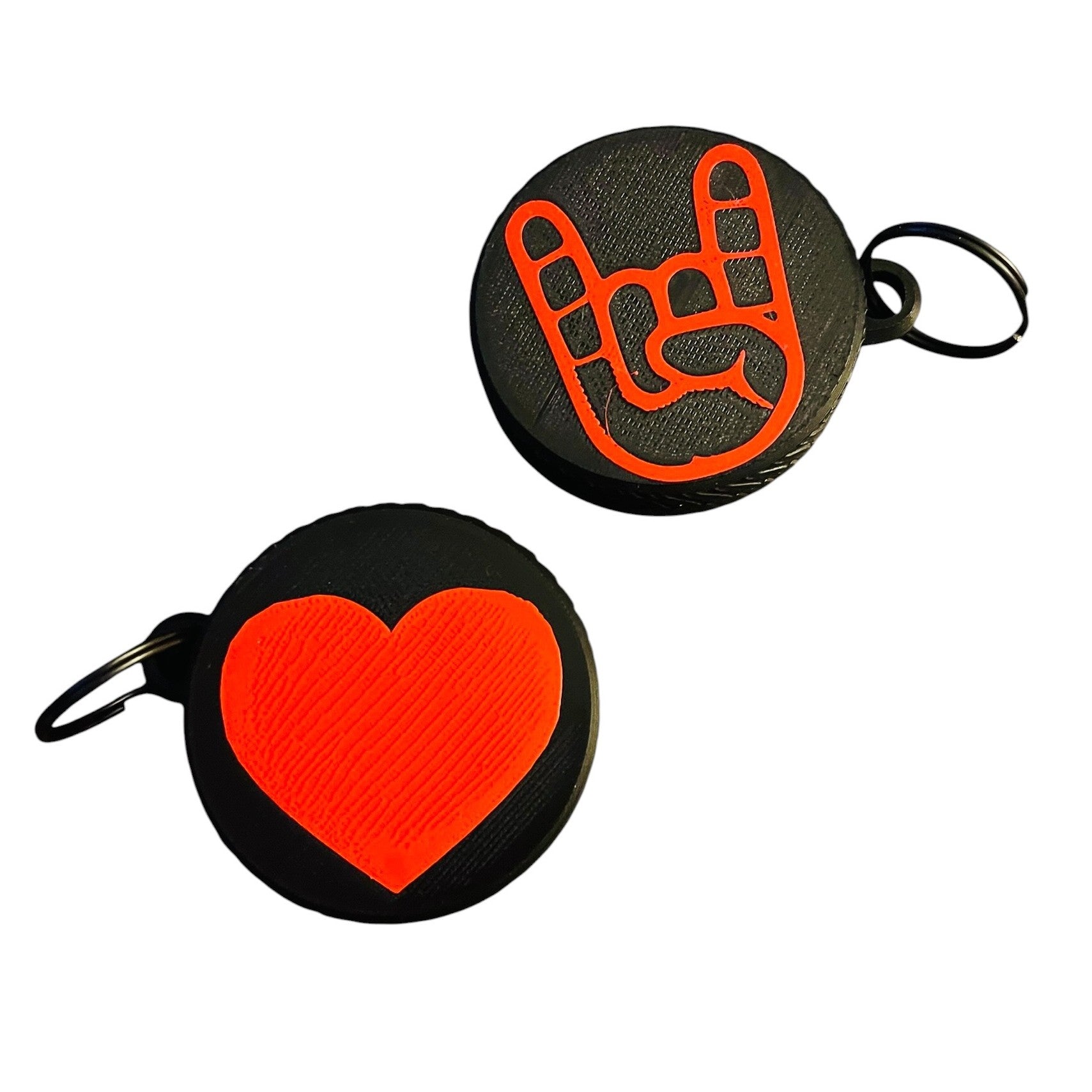 Two round black screw top container keychains on a white background, one featuring a bright red rock on gesture symbol and the other featuring a textured bright red heart. Both feature a black split ring.