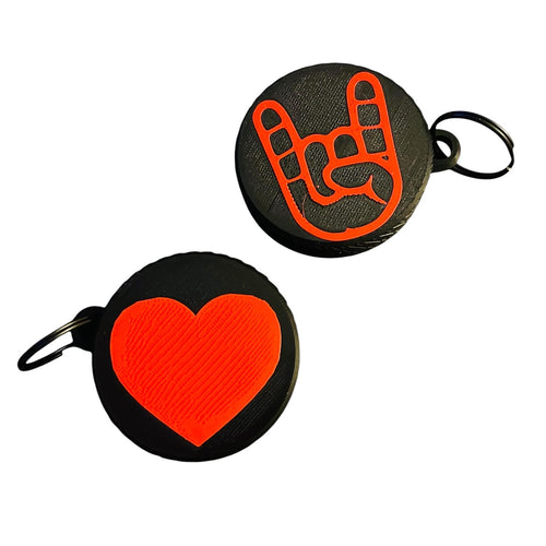 Two round black screw top container keychains on a white background, one featuring a bright red rock on gesture symbol and the other featuring a textured bright red heart. Both feature a black split ring.