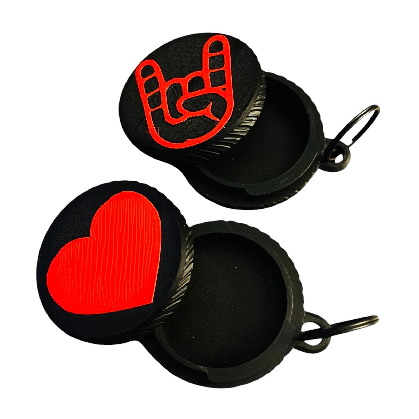 Two round black screw top container keychains on a white background, one featuring a bright red rock on gesture symbol and the other featuring a textured bright red heart. Both feature a black split ring.