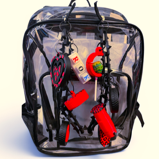 A clear festival backpack with black trim and handles, shown against a plain white background. Attached to the front are several novelty keychains and small accessories, including red cylindrical items, a white-and-black letter cube, and black spiky clips.