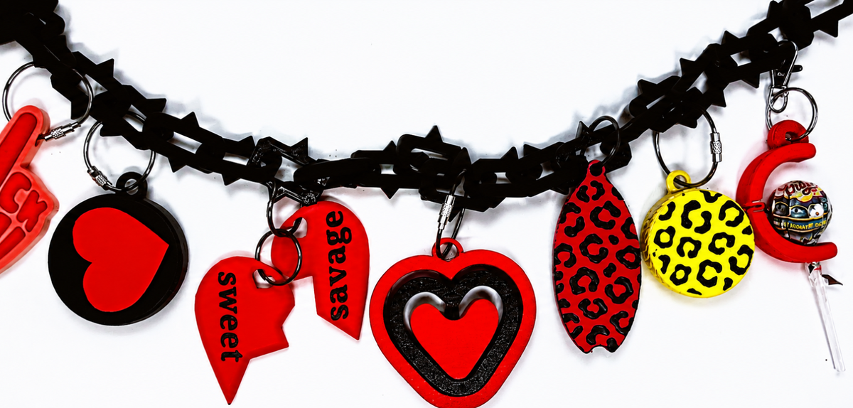 A close-up of a black bag chain adorned with multiple colorful charms, including red and black heart charms, a red and black leopard print charm, a yellow leopard print charm, and a red letter C charm with a lollipop.